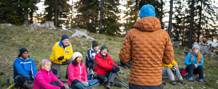 Kuvan etualalla selin kuvaajaan seisoo opas, jota kuuntelee joukko metsänrinteessä istuvia henkilöitä.