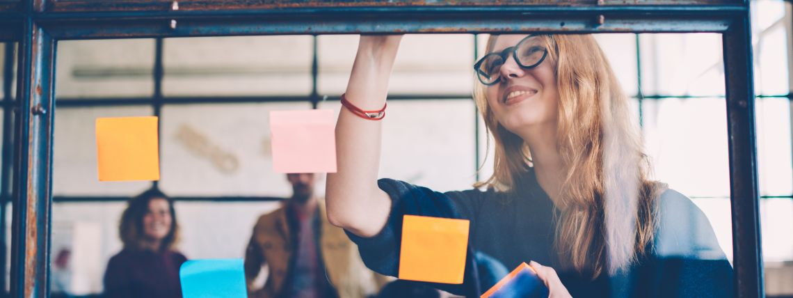 Korkeakouluopiskelijat katsovat kuinka toinen opiskelija järjestelee post-it lappuja lasiseinälle.