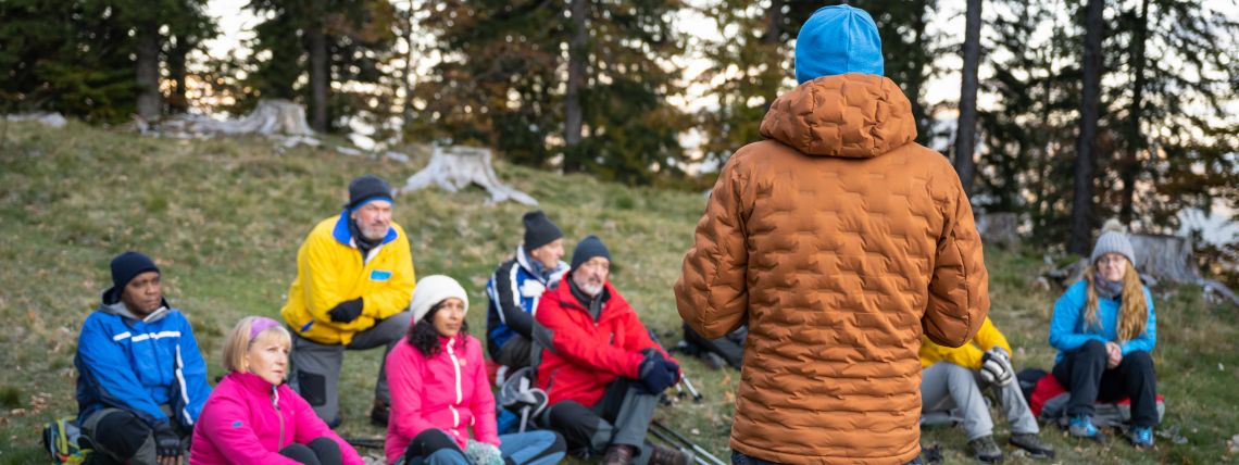 Kuvan etualalla selin kuvaajaan seisoo opas, jota kuuntelee joukko metsänrinteessä istuvia henkilöitä.